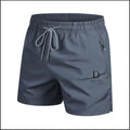 A pair of men's athletic shorts in slate gray, featuring a matching gray elastic waistband with a silver-and-white patterned drawstring, a side zip pocket, and a black 'Della of A' logo printed on the lower left leg.